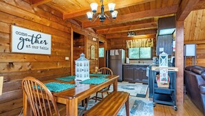Interior de una cabaña de madera con mesa de comedor, sillas, banco, zona de cocina y un espacio de sala acogedor.