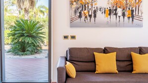 Een woonkamer met een bruine bank en gele kussens, een schilderij van mensen op een brug, en een grote plant bij de deur.