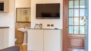 Een kleine hotelkamer met een flatscreen televisie boven de wastafel, een houten deur met glaspanelen en een tafeltje met een stoel.