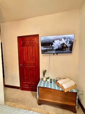 A room with a wooden door, a television mounted on the wall displaying a weather forecast, a small table with a blue and white striped tablecloth, and a wooden bench.