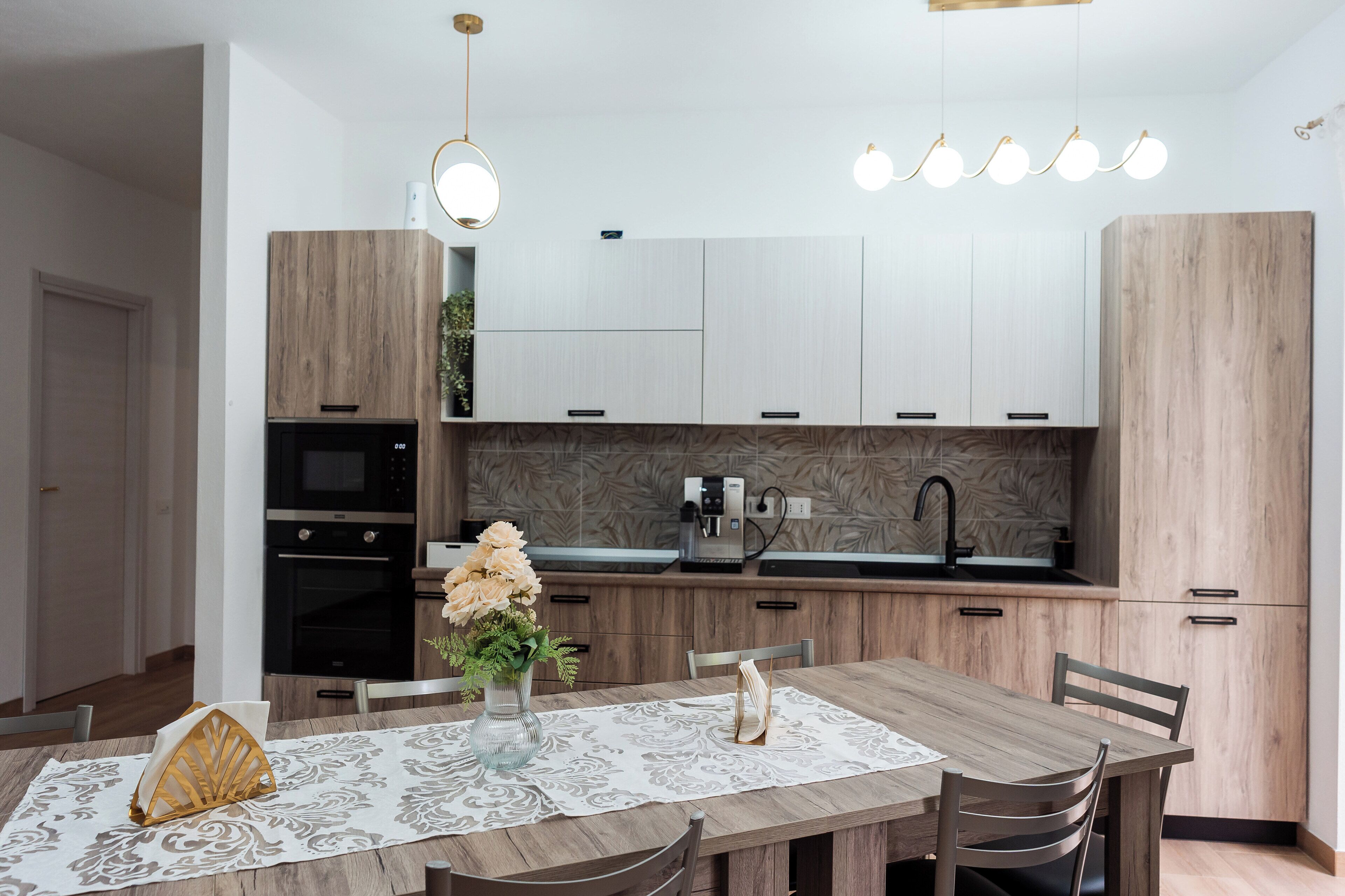 A modern kitchen with wooden cabinets, a built-in oven, a coffee machine, and a dining area with a table and chairs.