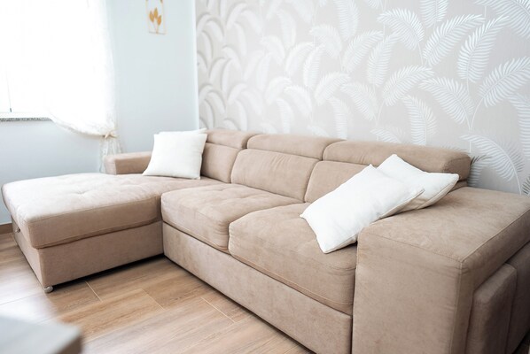 Un sofá seccional color beige con cojines blancos en una habitación con piso de madera y papel tapiz estampado con helechos.