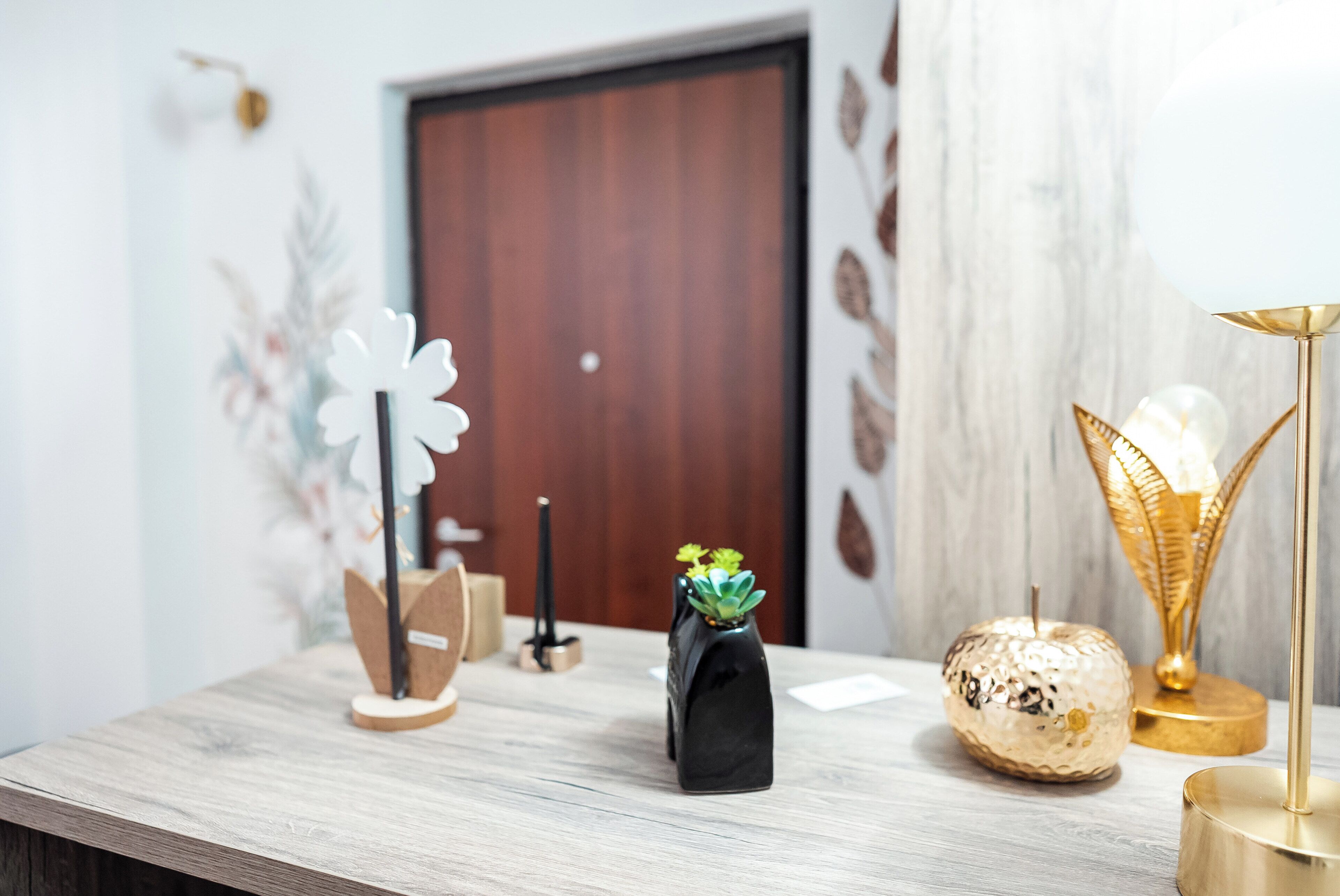 Una mesa de madera con objetos decorativos, incluyendo un jarrón negro con una suculenta, un objeto esférico dorado y una lámpara dorada.