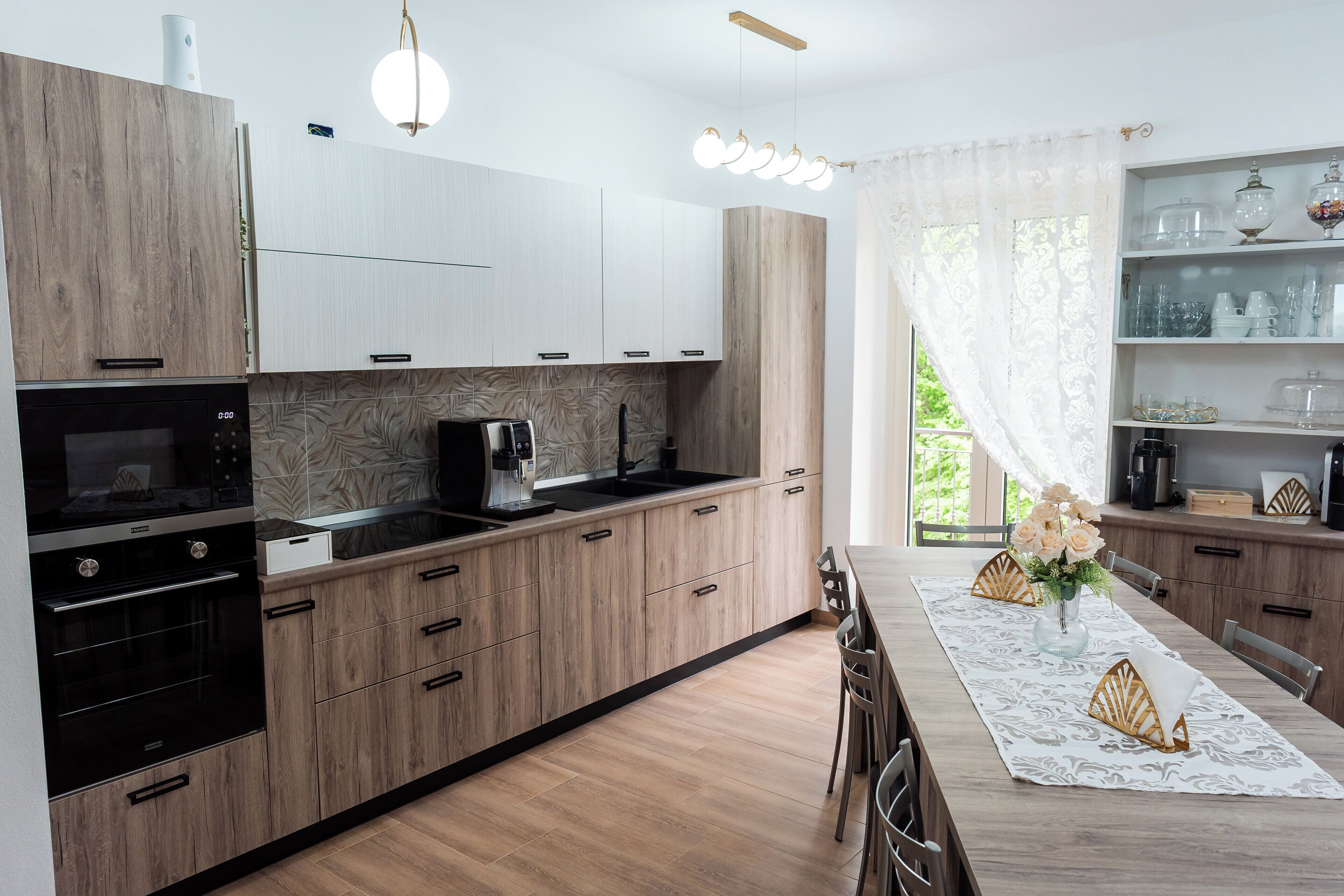 A modern kitchen with wooden cabinets, a built-in oven, a coffee machine, and a dining area with a table set for two.