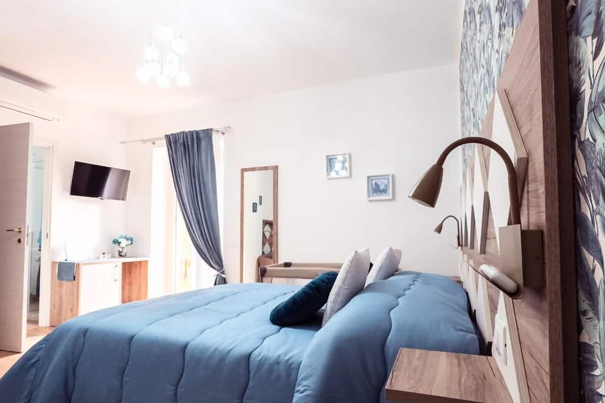 Et moderne soveværelse med en træseng, et blåt quiltet dynebetræk, et natbord med en lampe og et vægmonteret fjernsyn.