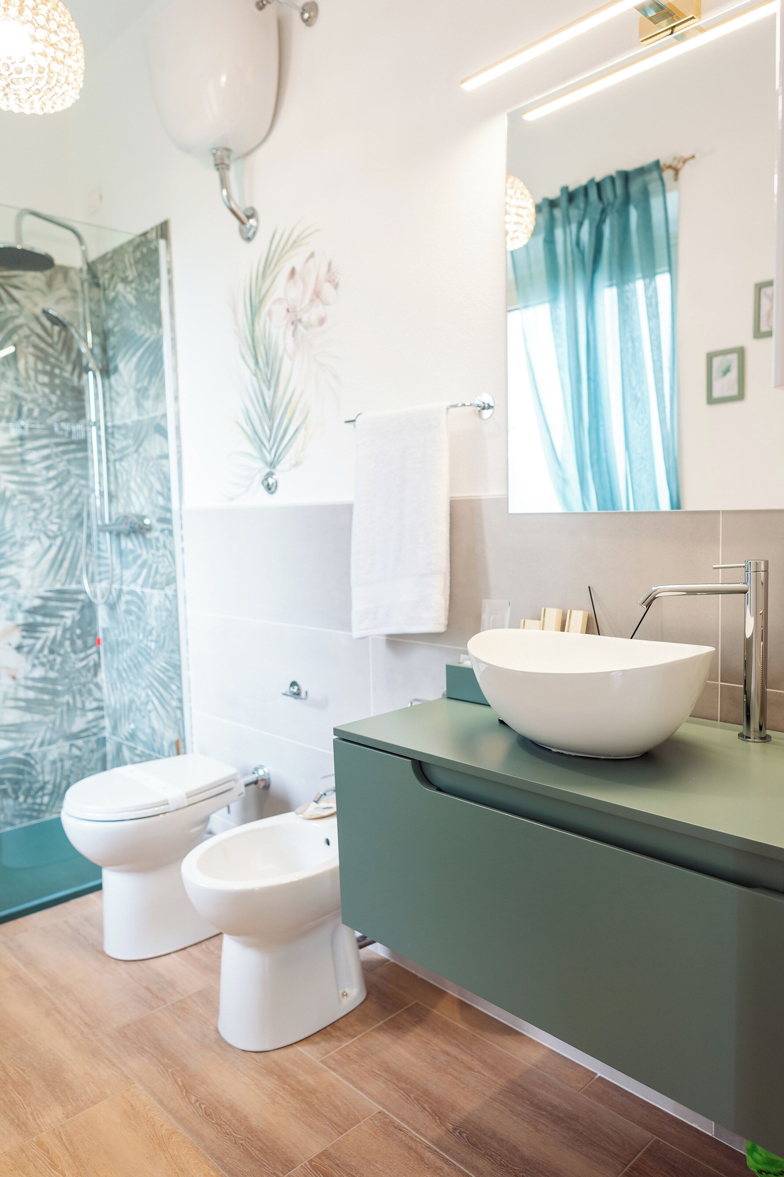 A modern bathroom with a large white vessel sink, two toilets, a glass shower enclosure, and a mirror with a blue curtain.