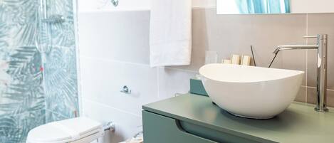 A modern bathroom with a large white vessel sink, two toilets, a glass shower enclosure, and a mirror with a blue curtain.