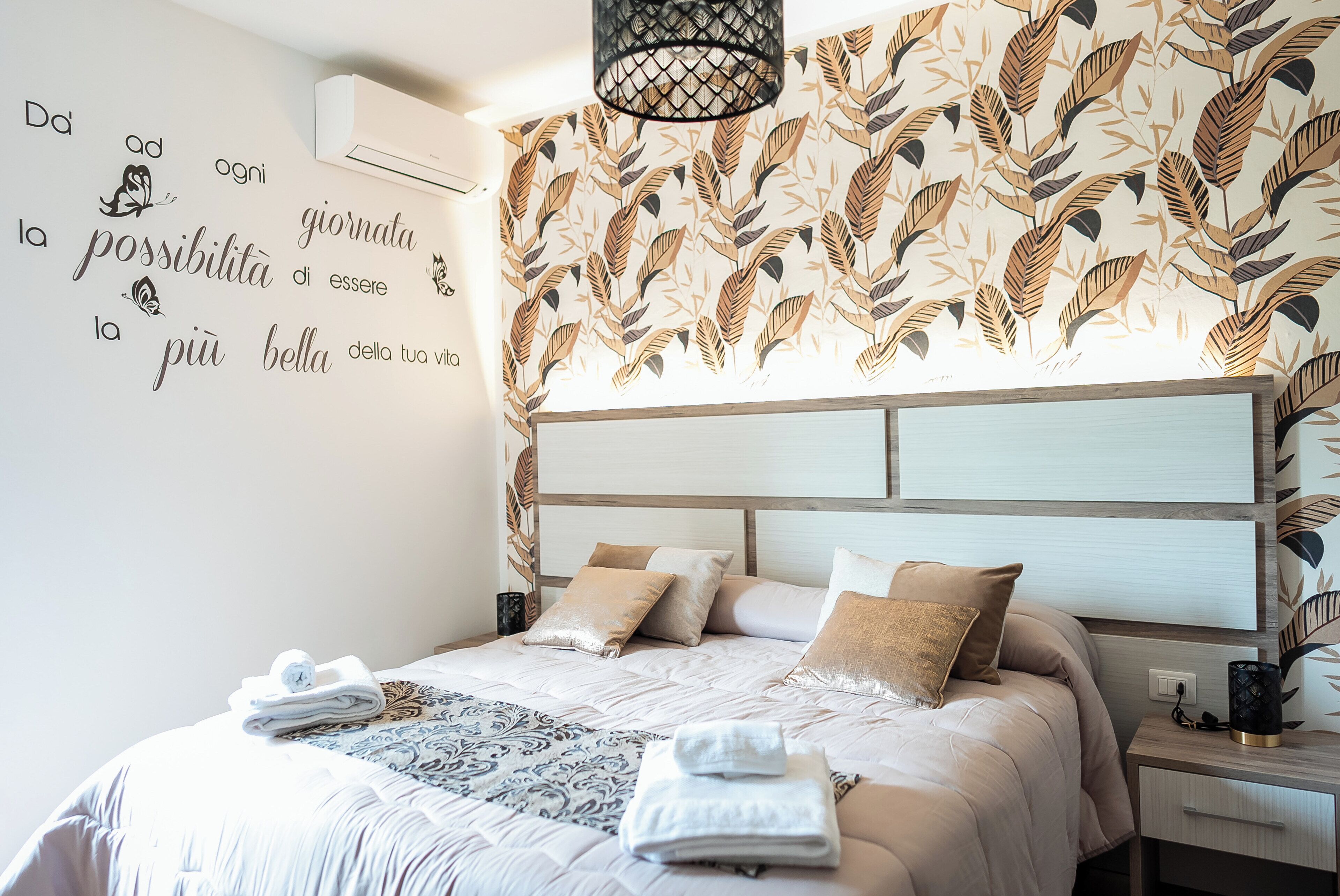 Un dormitorio con cama, almohadas, una pared con diseño de hojas, un equipo de aire acondicionado y una mesita de noche.