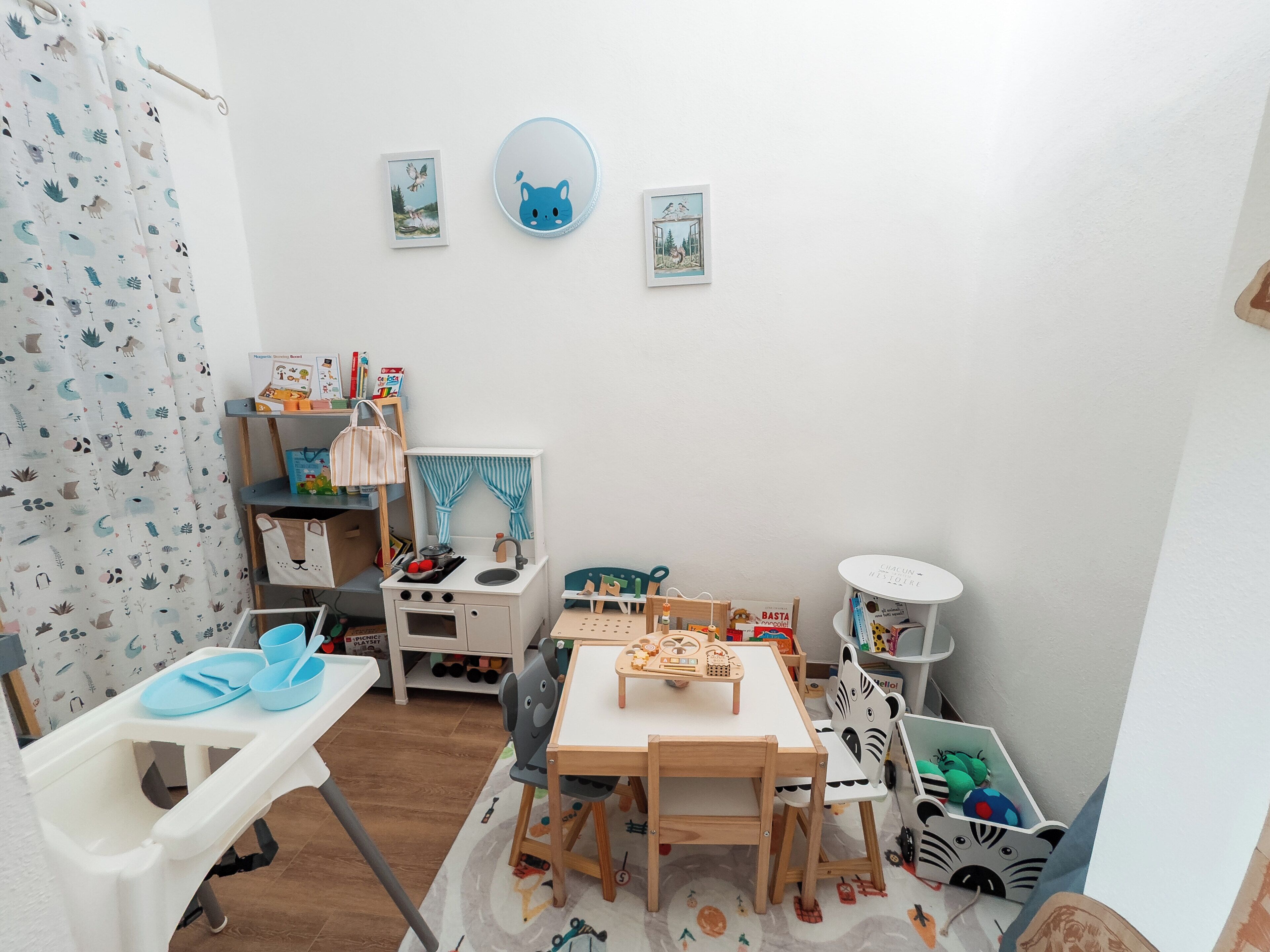 Una stanza giochi per bambini con un tavolino, sedie, una libreria e un angolo cottura giocattolo.