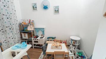 Una stanza giochi per bambini con un tavolino, sedie, una libreria e un angolo cottura giocattolo.