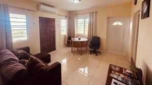 Una habitación con un sofá, una mesa pequeña con una silla, una puerta y ventanas con cortinas.