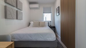 A bedroom with a bed, bedside tables, an air conditioning unit, a window, and a wooden panel on the wall.