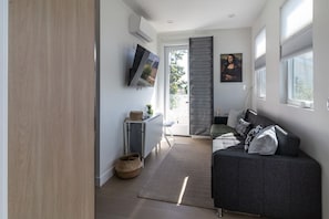 A modern living room with a grey sofa, a flat-screen TV mounted on the wall, and a large window with blinds.