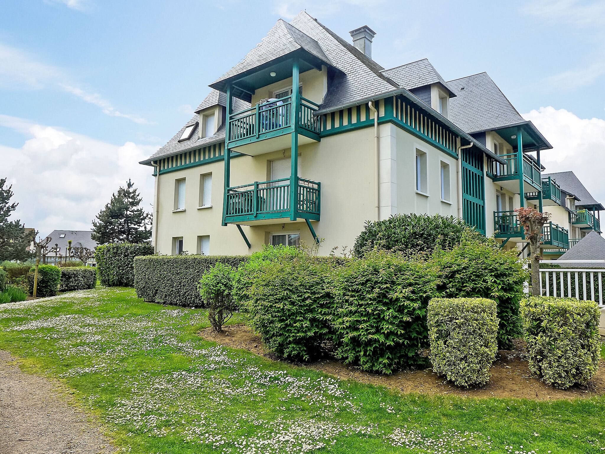 Un bâtiment à deux étages doté d’un balcon, entouré de haies bien entretenues et d’une pelouse agrémentée de fleurs blanches.