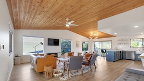A spacious living area with a wooden ceiling, a dining table, and a kitchen area with blue cabinets. - Surf Shack at Lake Nacimiento with Private Slip! (Bradley)