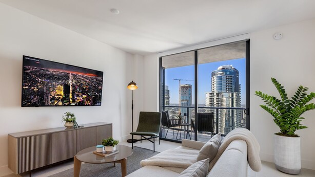A modern living room with a large window, a sofa, a coffee table, a TV, and a potted plant. - 601 RESIDENCES SKY FLOOR 1BR UNIT #4511 (Miami)
