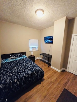 A bedroom with a bed, a TV mounted on the wall, a small table, and a window with blinds.