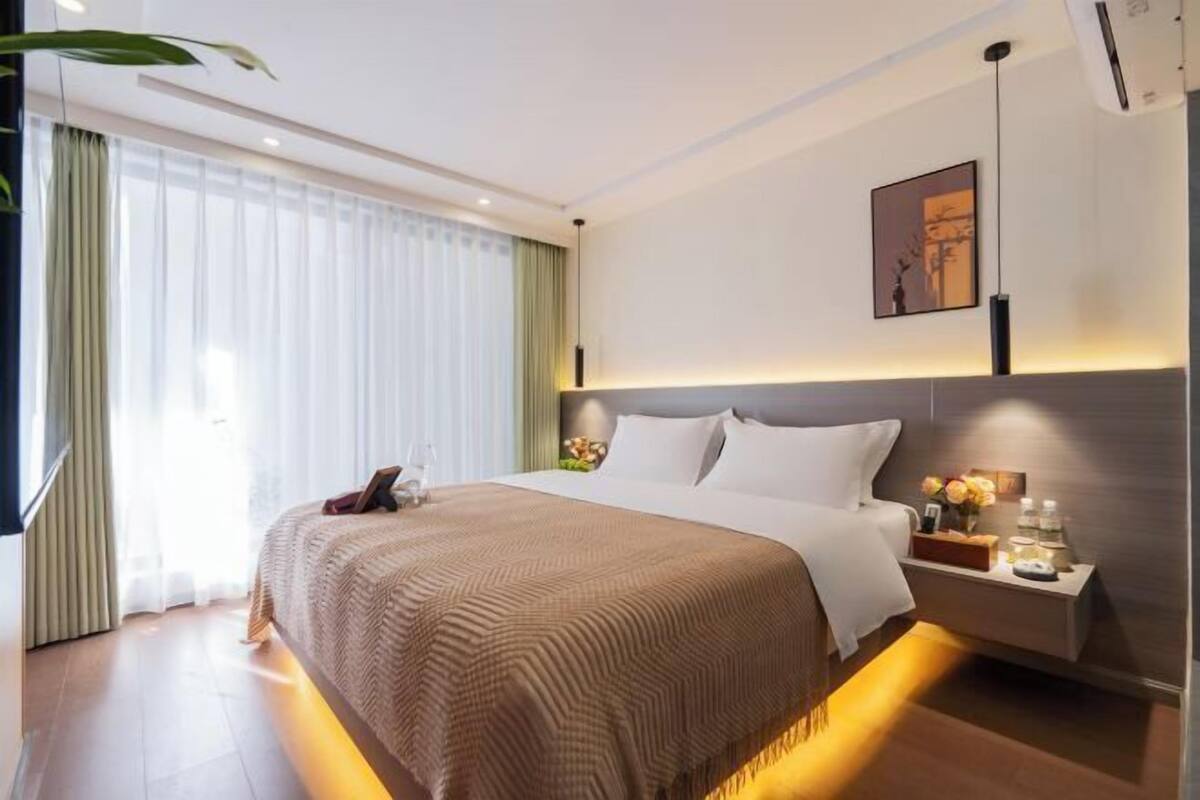 Une chambre d’hôtel moderne avec un grand lit, une table de chevet avec une lampe et une vue sur la ville grâce à de grandes fenêtres.