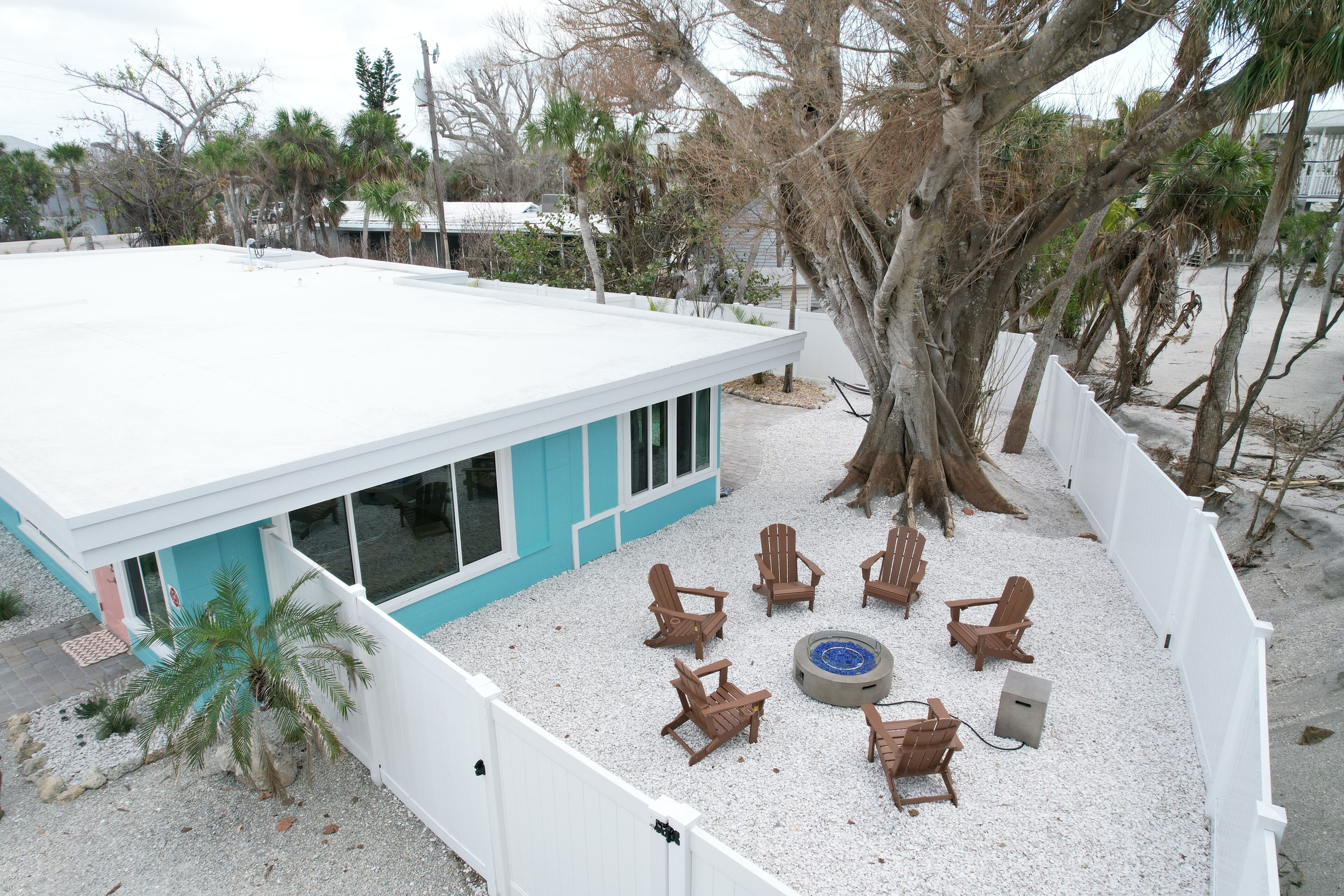 A modern house with a white roof and turquoise accents, surrounded by a white fence and featuring a patio with wooden chairs and a fire pit.