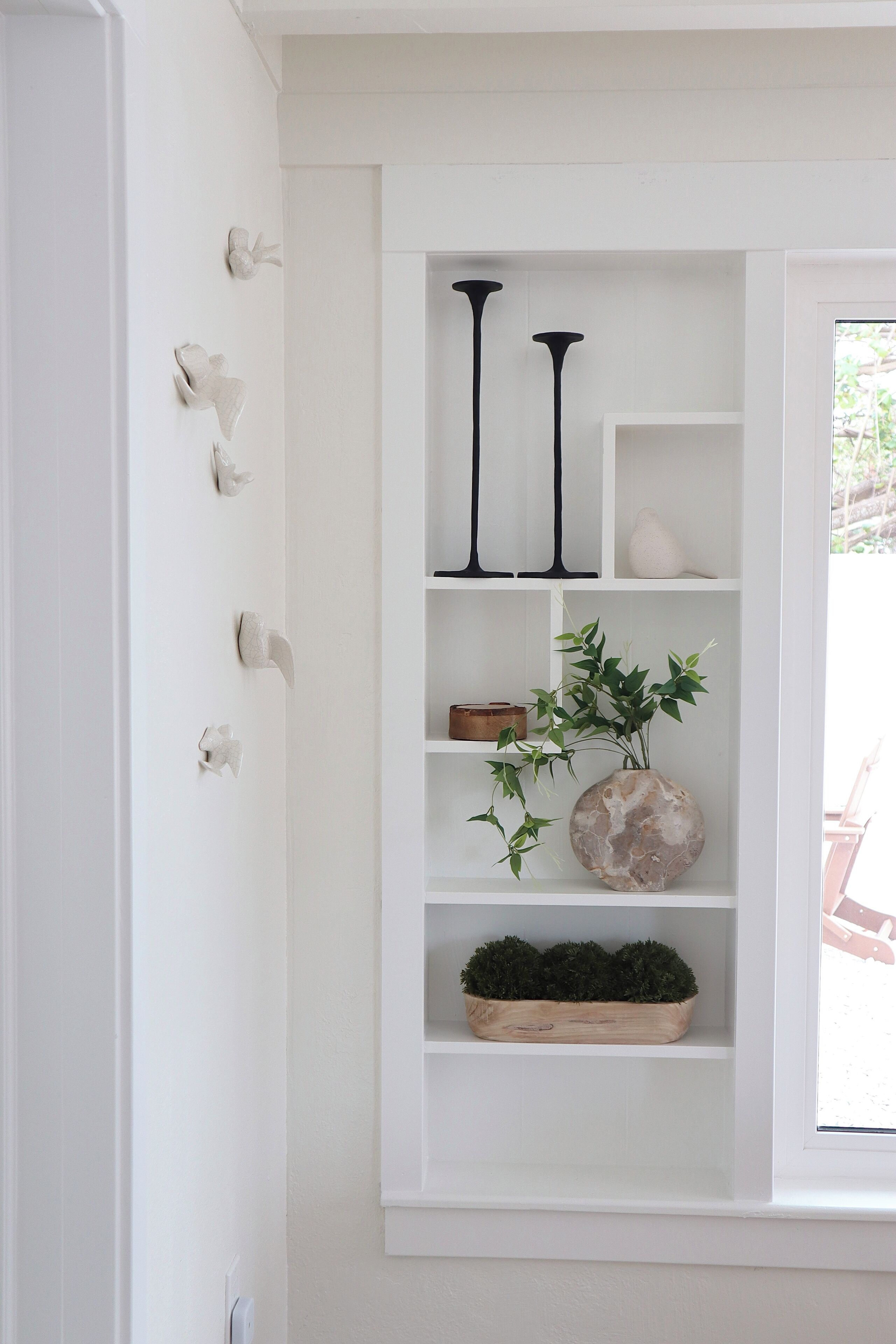 A white shelf unit with decorative items including black candlesticks, a vase with green foliage, and a wooden box.