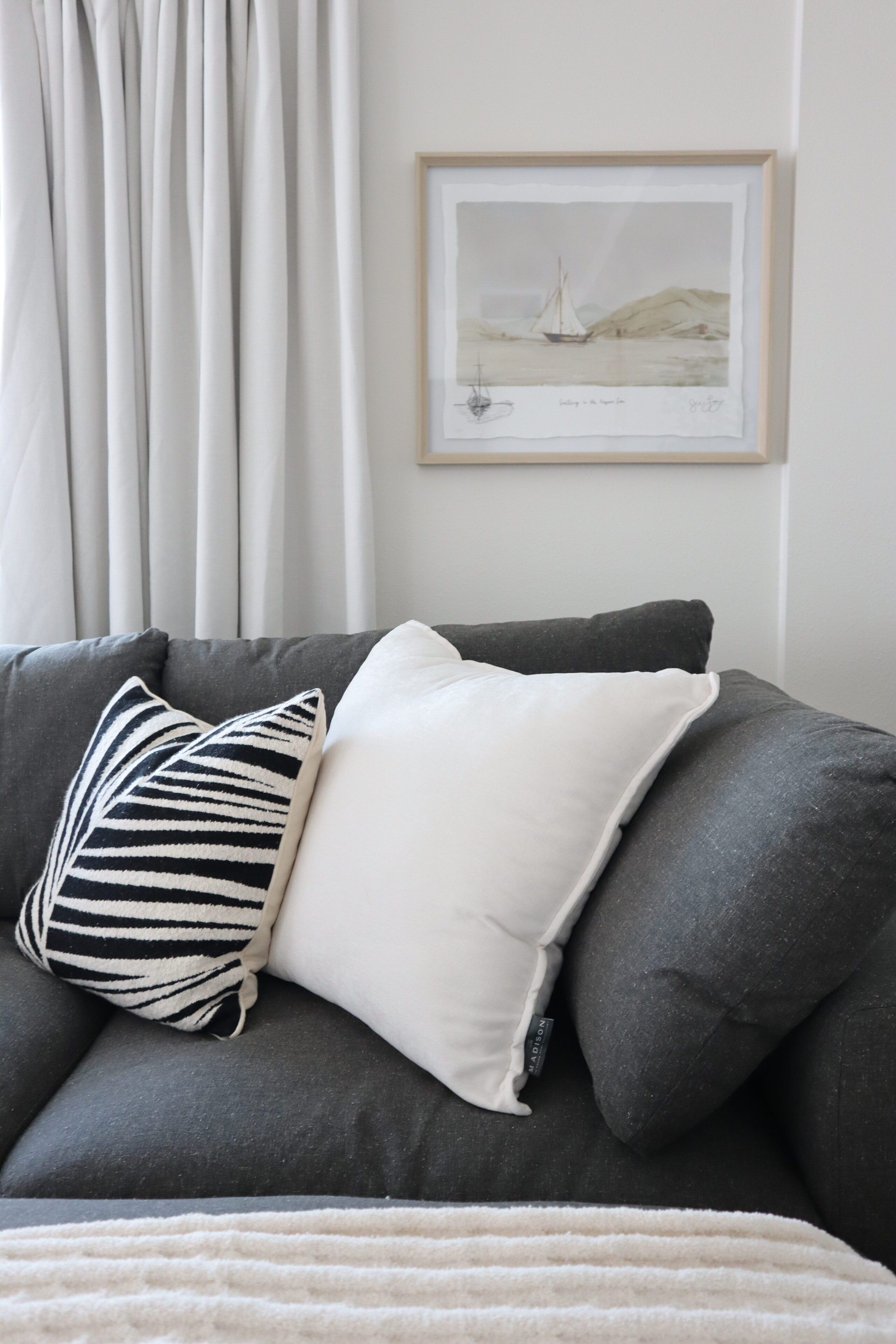 Un canapé gris agrémenté d’un coussin blanc et d’un coussin à rayures, une œuvre d’art encadrée représentant un voilier, et un rideau blanc.