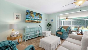 Ein Wohnzimmer mit Blick auf einen Pool, ein Korbstuhl, ein weißes Sofa, ein Glastisch und ein Gemälde mit einer Strandansicht.