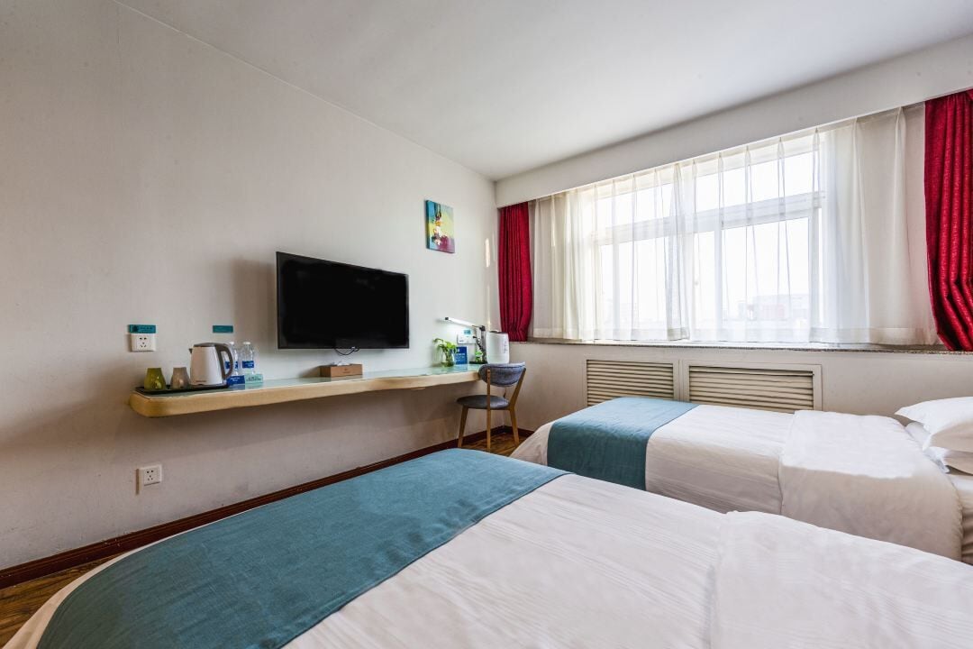 Ett hotellrum med två sängar, en tv som är monterad på väggen, ett litet skrivbord med en stol och ett fönster med röda gardiner.