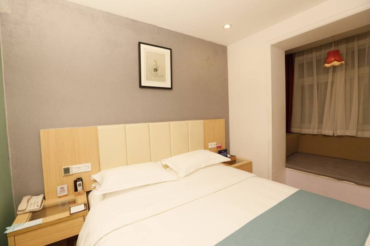A hotel room with a bed, a bedside table with a phone, a digital clock, and a window with a sheer curtain.