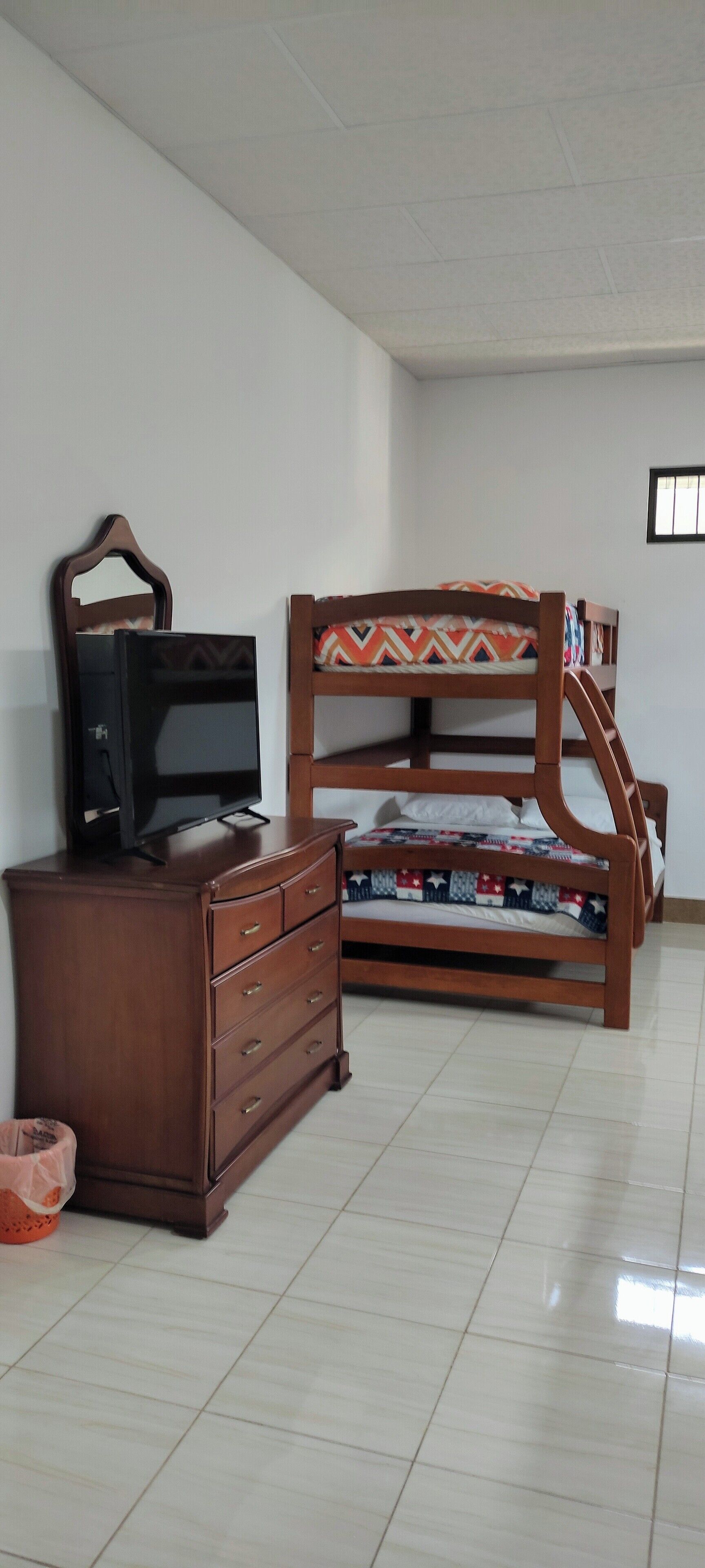A room with a wooden bed with a patterned bedspread, a wooden dresser with a television on top, and a tiled floor.