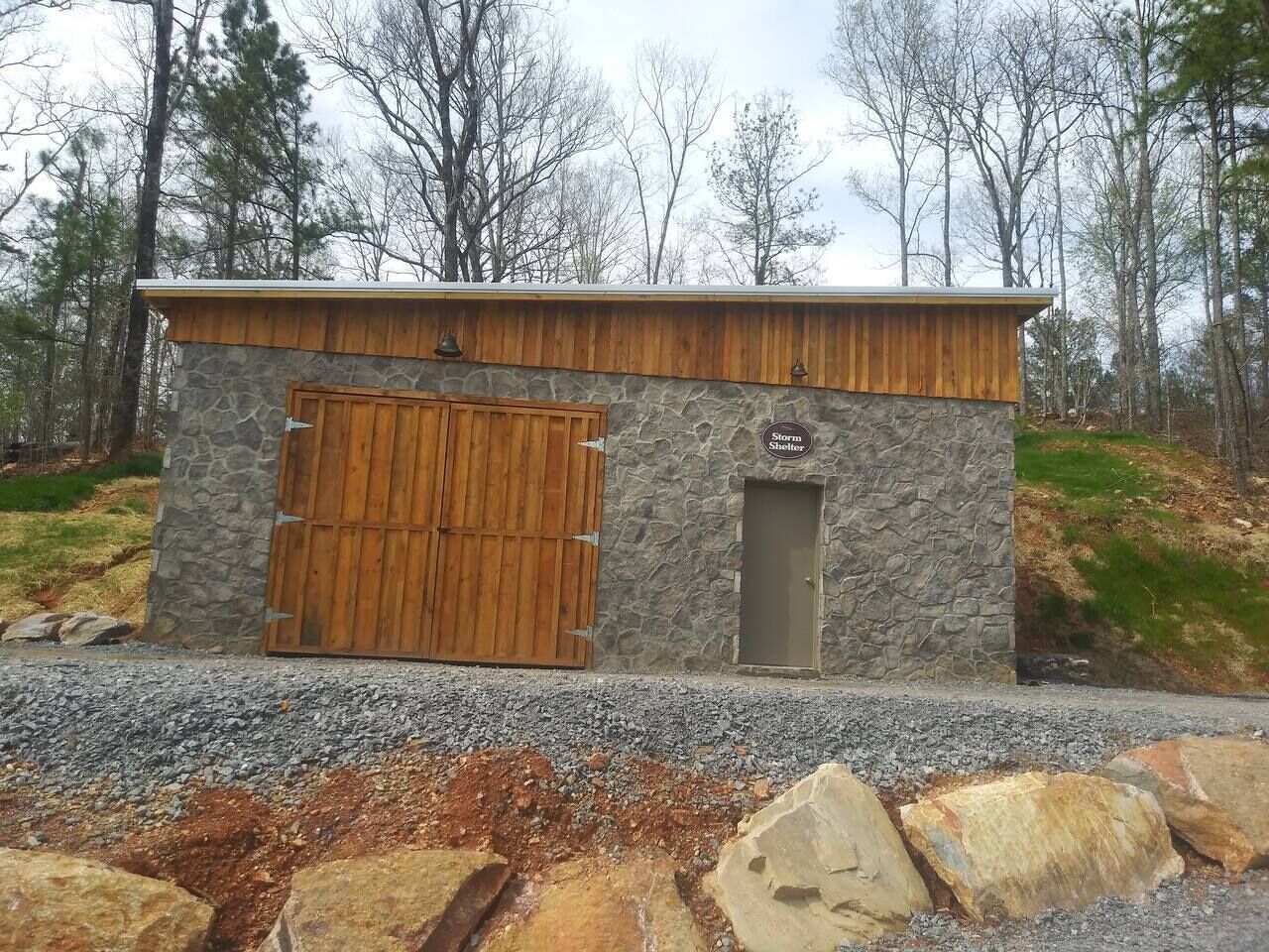 A stone building with a wooden door and a small window, surrounded by trees and gravel.