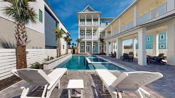 A pool area with lounge chairs, a pool, and a two-story building with balconies. - Deja View - Ocean Views for Miles with Private Pool, Hot Tub, Game Room & Easy Beach Access (Port Aransas)