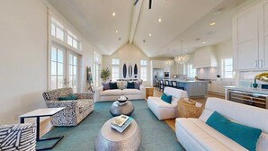 A spacious living area with a high ceiling, large windows, and a modern kitchen. - Deja View - Ocean Views for Miles with Private Pool, Hot Tub, Game Room & Easy Beach Access (Port Aransas)