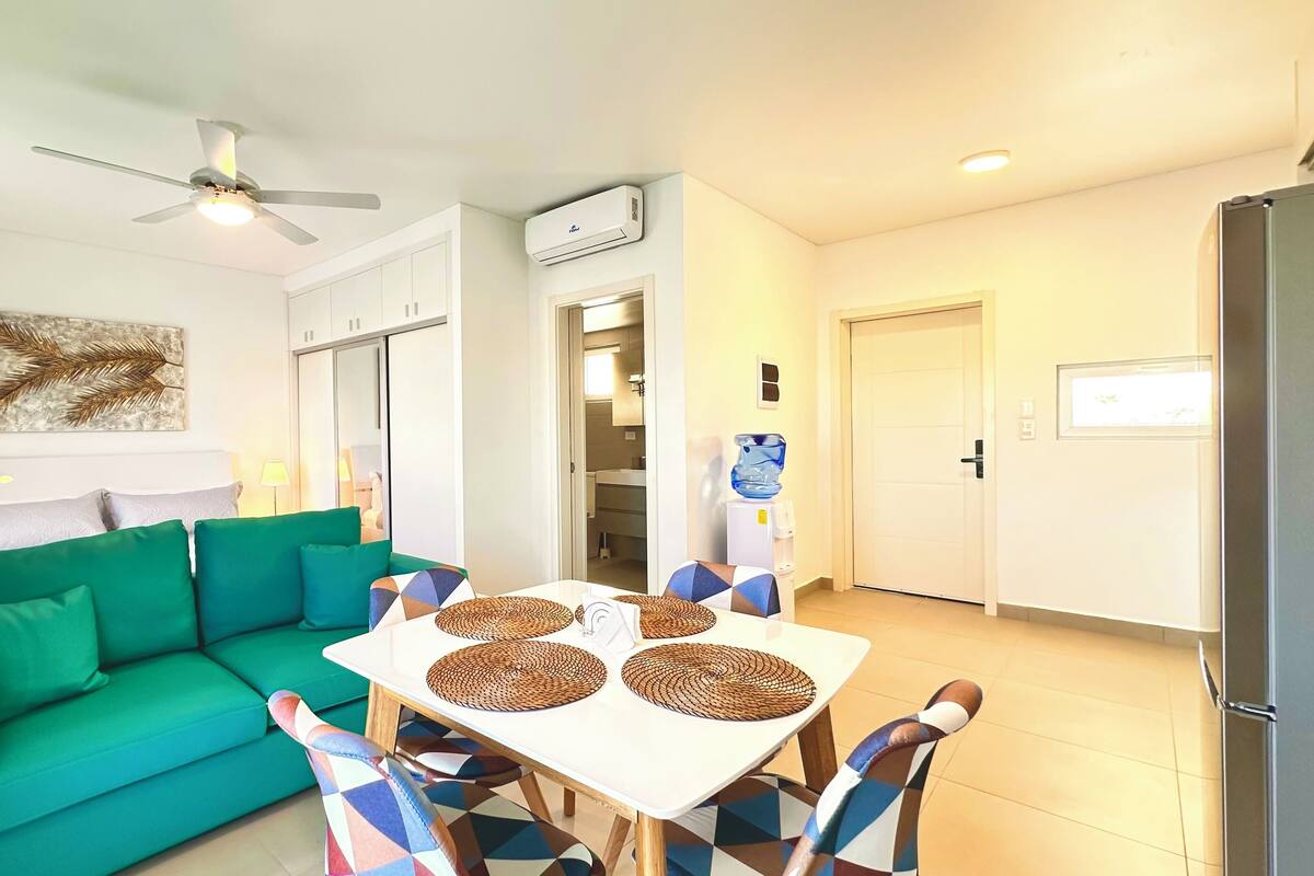 A modern living room with a green sofa, a dining table with woven placemats, a ceiling fan, and a water dispenser.