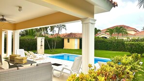 A covered outdoor area with a pool, lounge chairs, and a small building in the background.