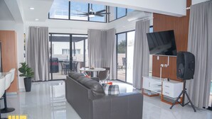 A modern living room with a grey sofa, a flat-screen TV, and a dining area with a table and chairs. - Rheia Properties - Sosua Ocean Village (Sosúa)