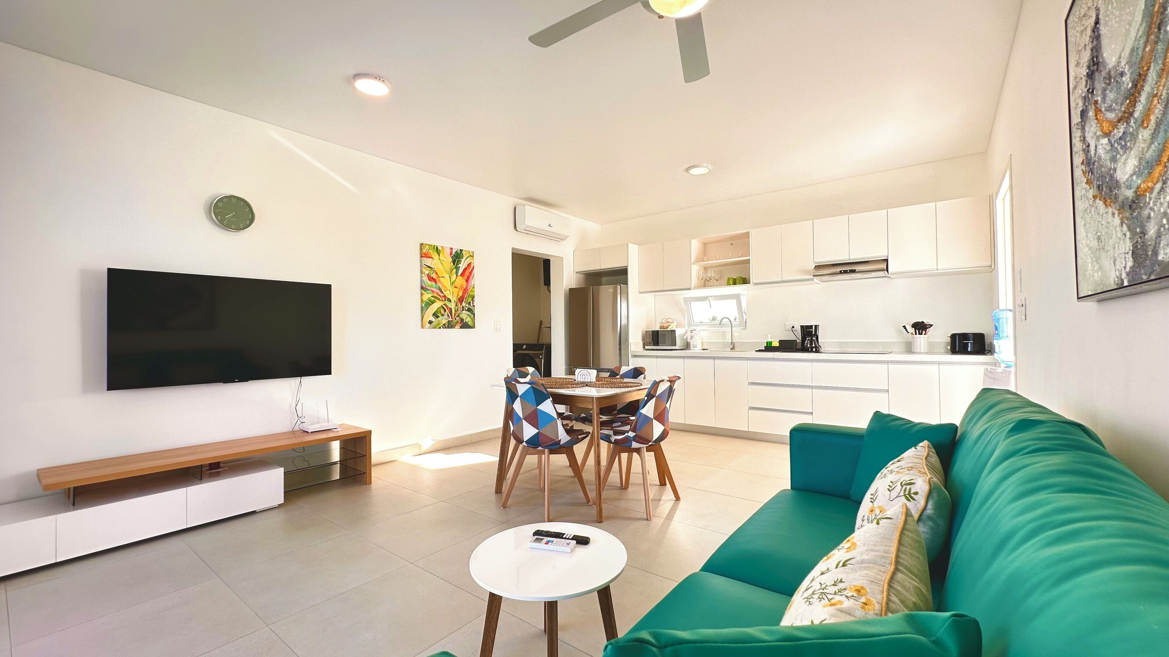 A modern living room with a flat-screen TV, a green sofa, a dining table with chairs, and a kitchen area in the background.