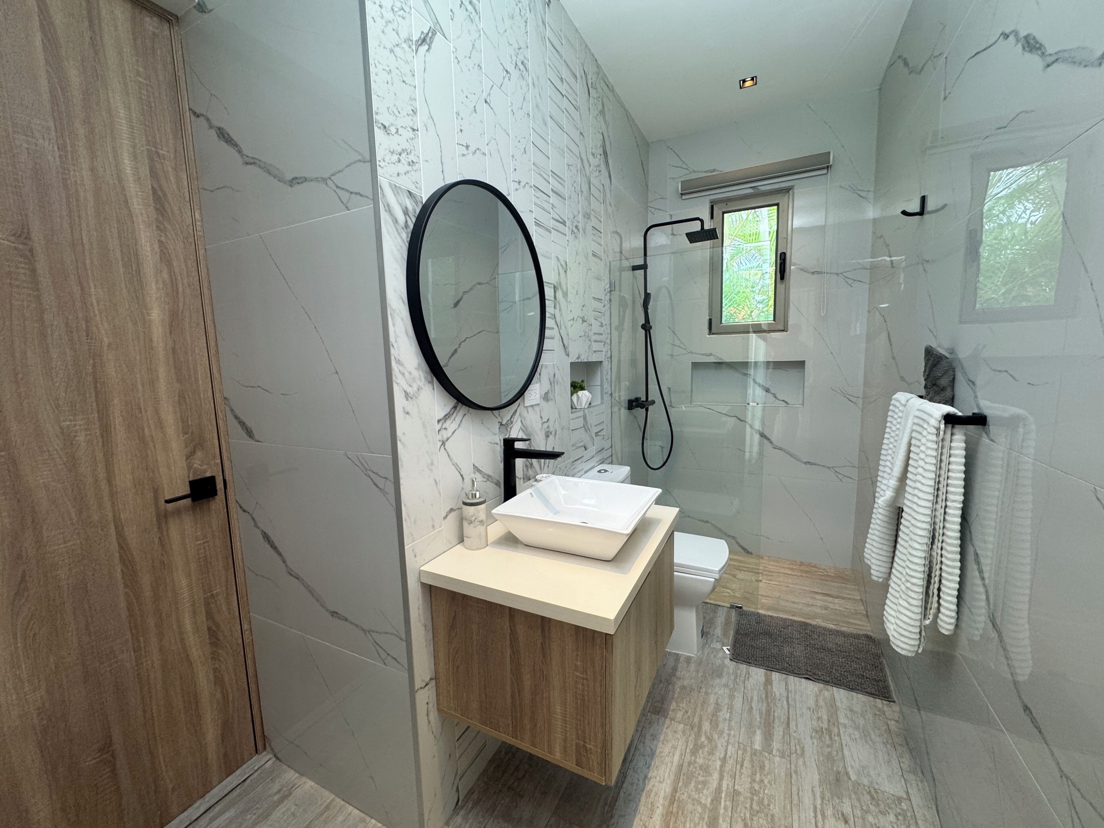 A modern bathroom with a marble wall, a wooden vanity, a round mirror, a shower, and a towel rack.