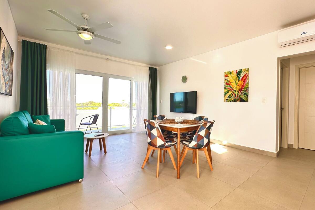A living room with a green sofa, a dining table with chairs, a ceiling fan, a TV, and a door.