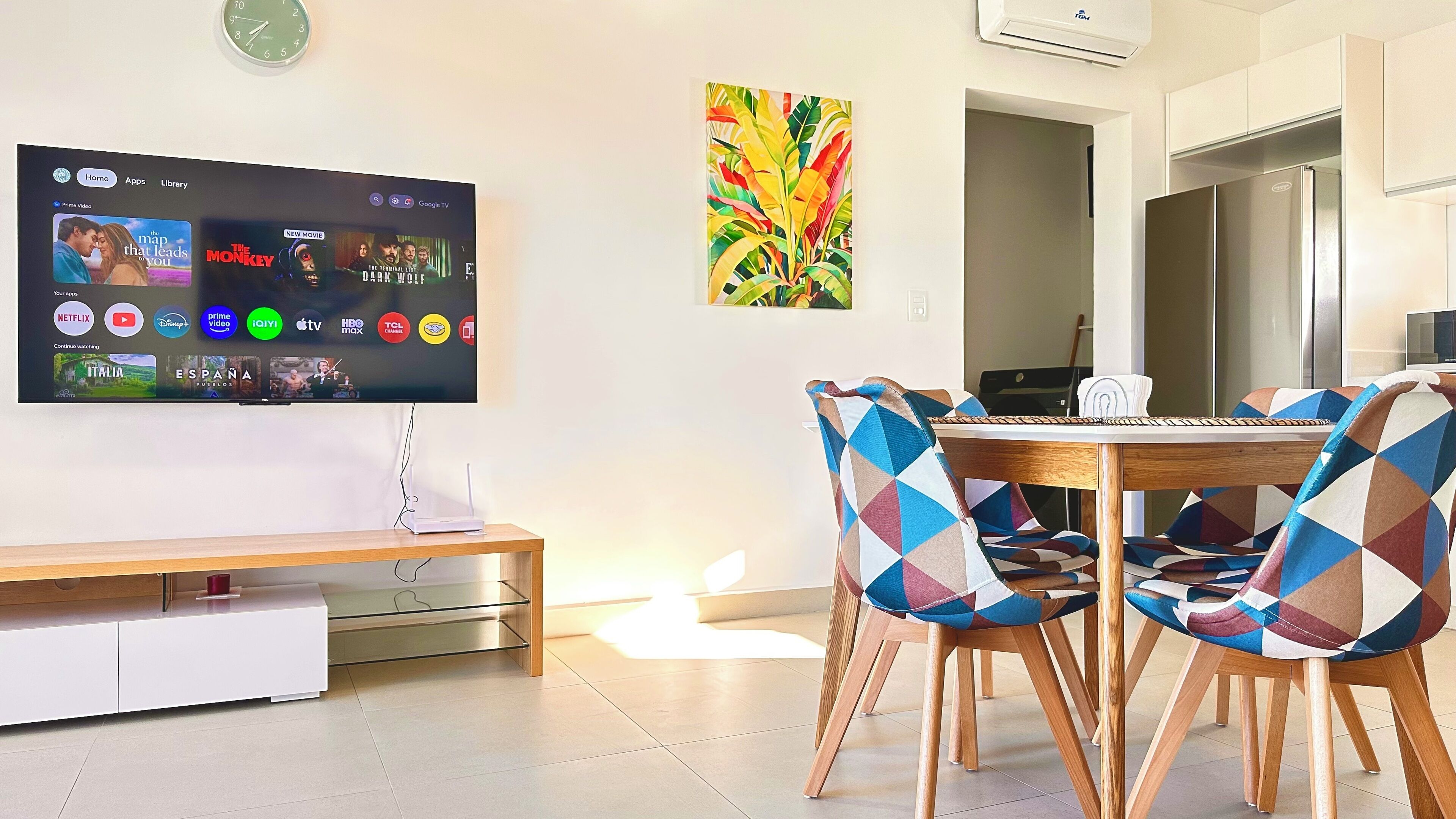 Ett modernt vardagsrum med en platt-TV som visar streamingtjänster, en matplats med mönstrade stolar och ett kök i bakgrunden.