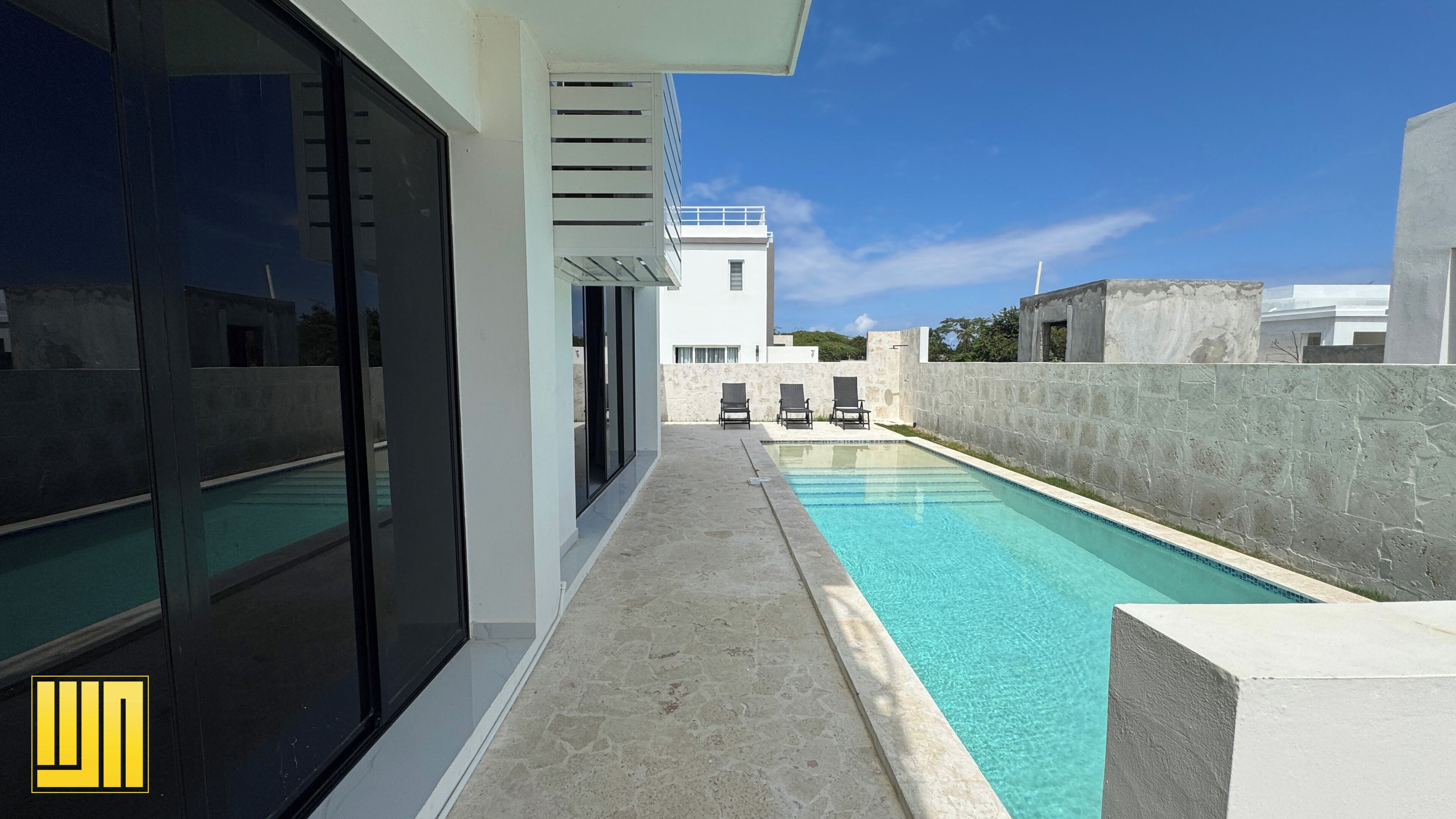 A modern pool area with a clear blue pool, a stone wall, and a white building with a balcony in the background.