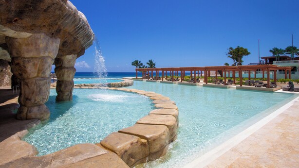 A pool with clear water, a rock formation, and a wooden pavilion with lounge chairs. - Rheia Properties - Sosua Ocean Village (Sosúa)