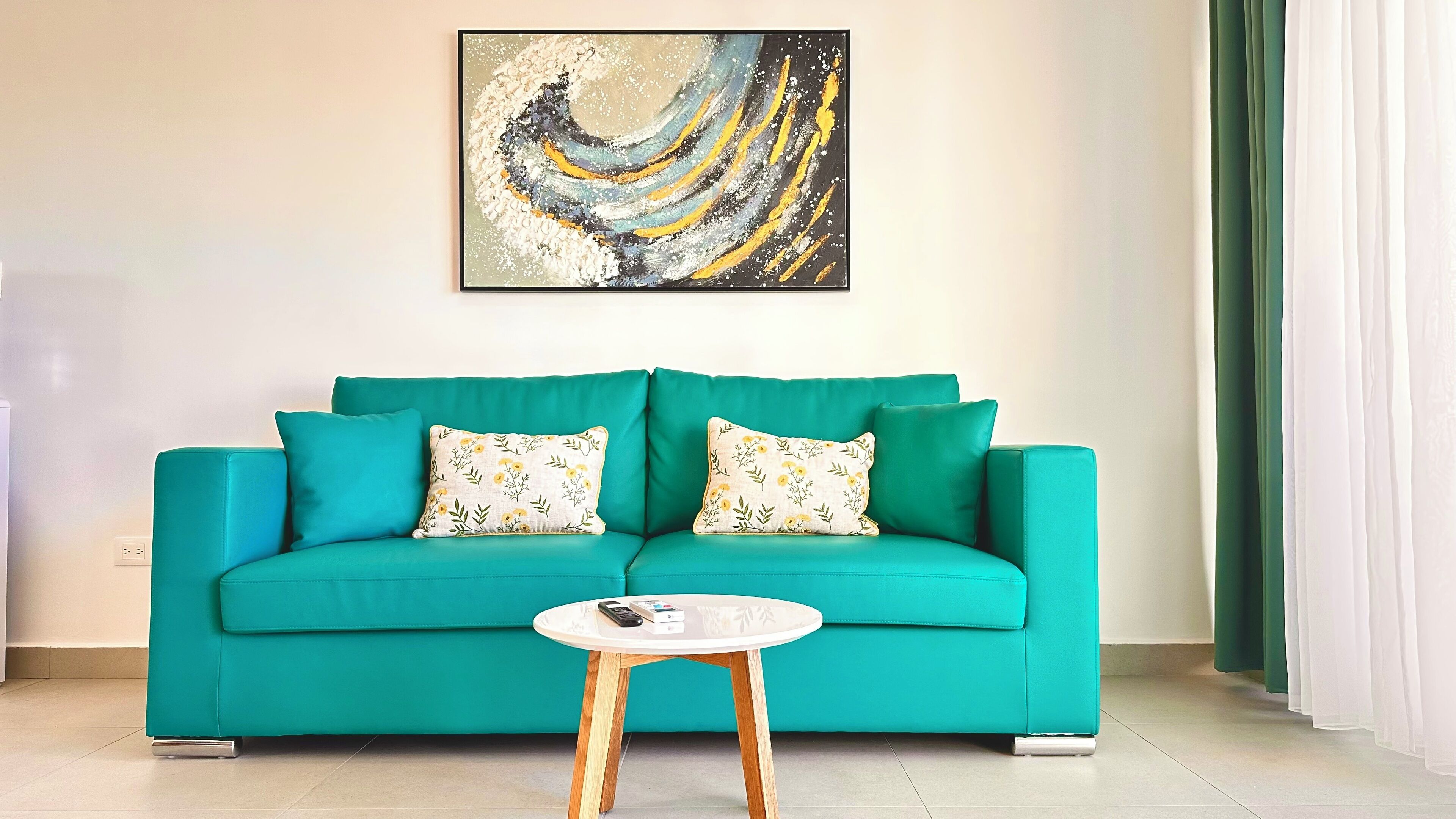Un canapé de couleur turquoise agrémenté de coussins à motifs floraux, une petite table ronde blanche avec une télécommande, et un tableau abstrait moderne accroché au mur.