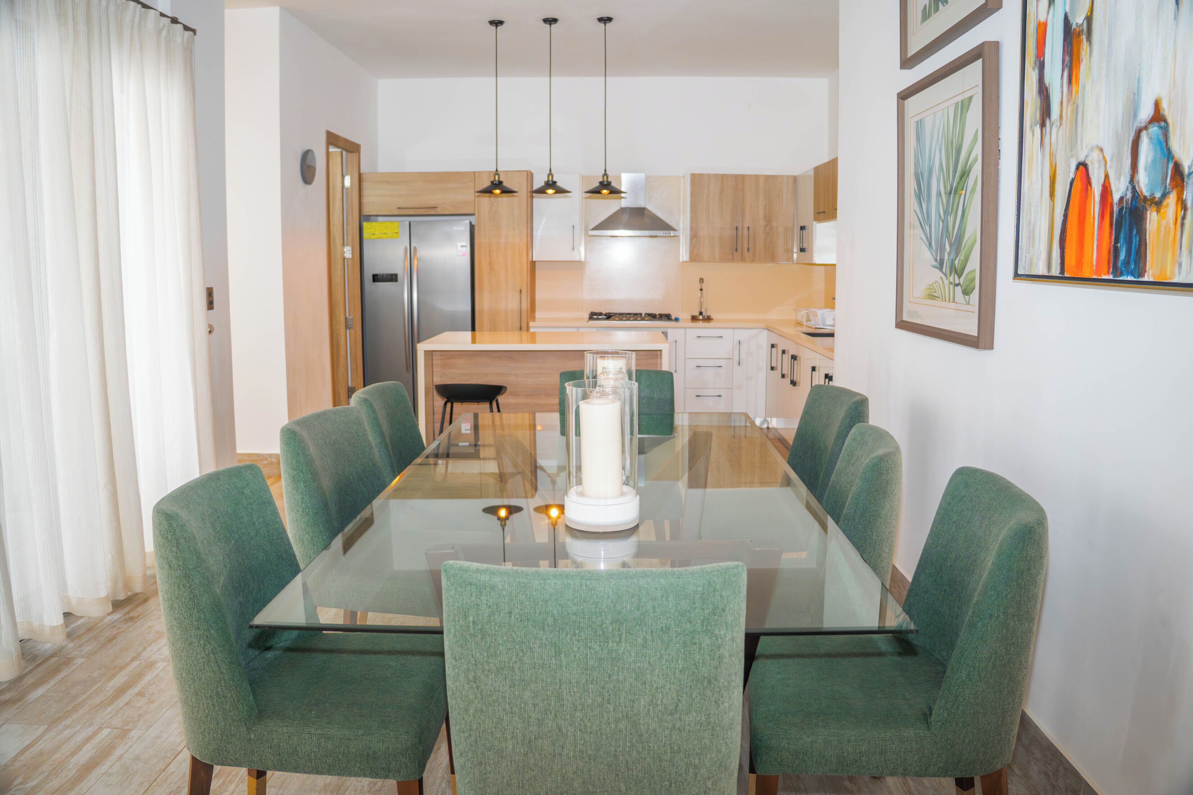 A modern dining area with a glass table, green upholstered chairs, a central candle, and a kitchen in the background.
