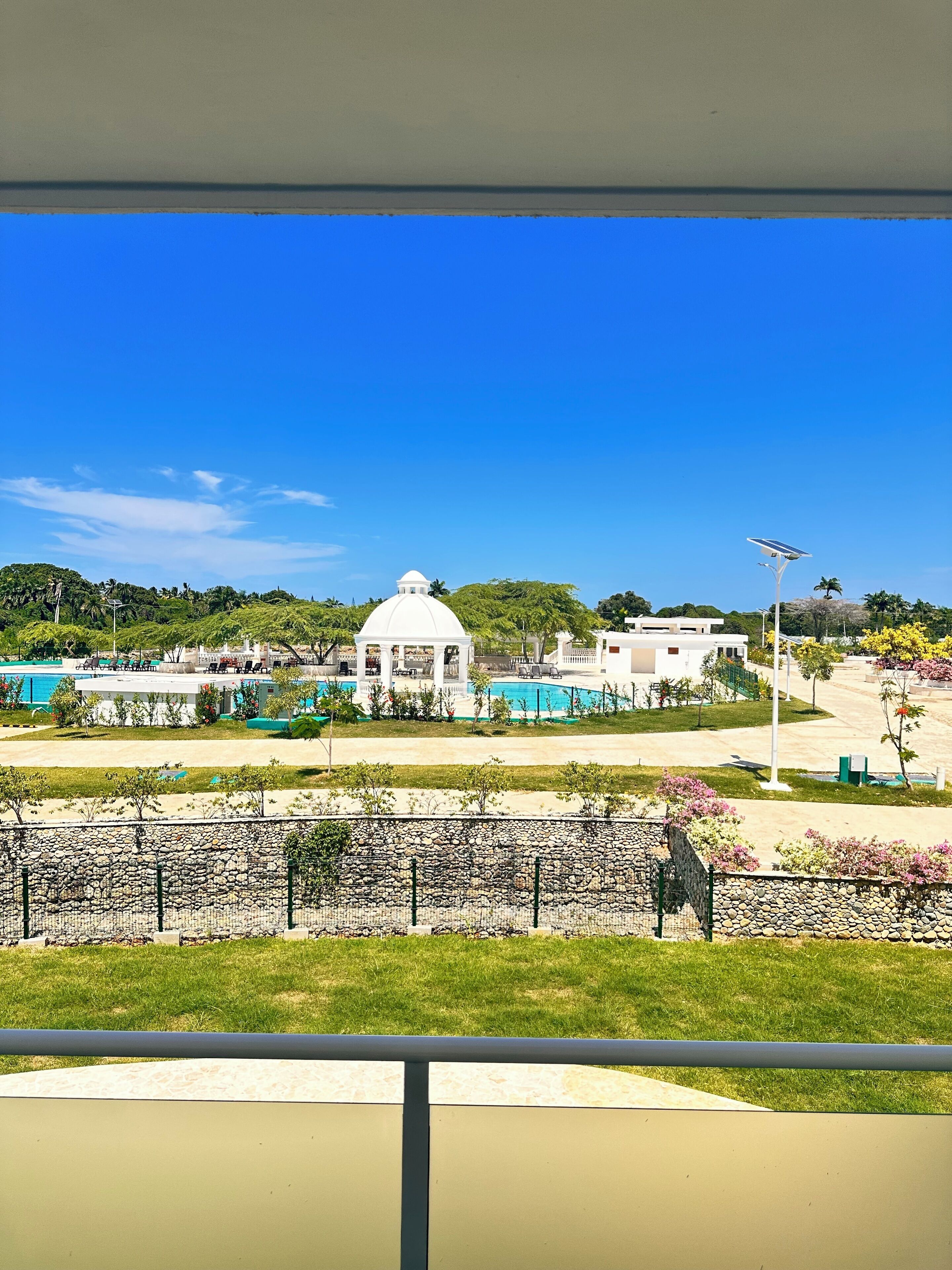 A view of a swimming pool area with a white gazebo and a dome structure, surrounded by greenery and a stone wall.