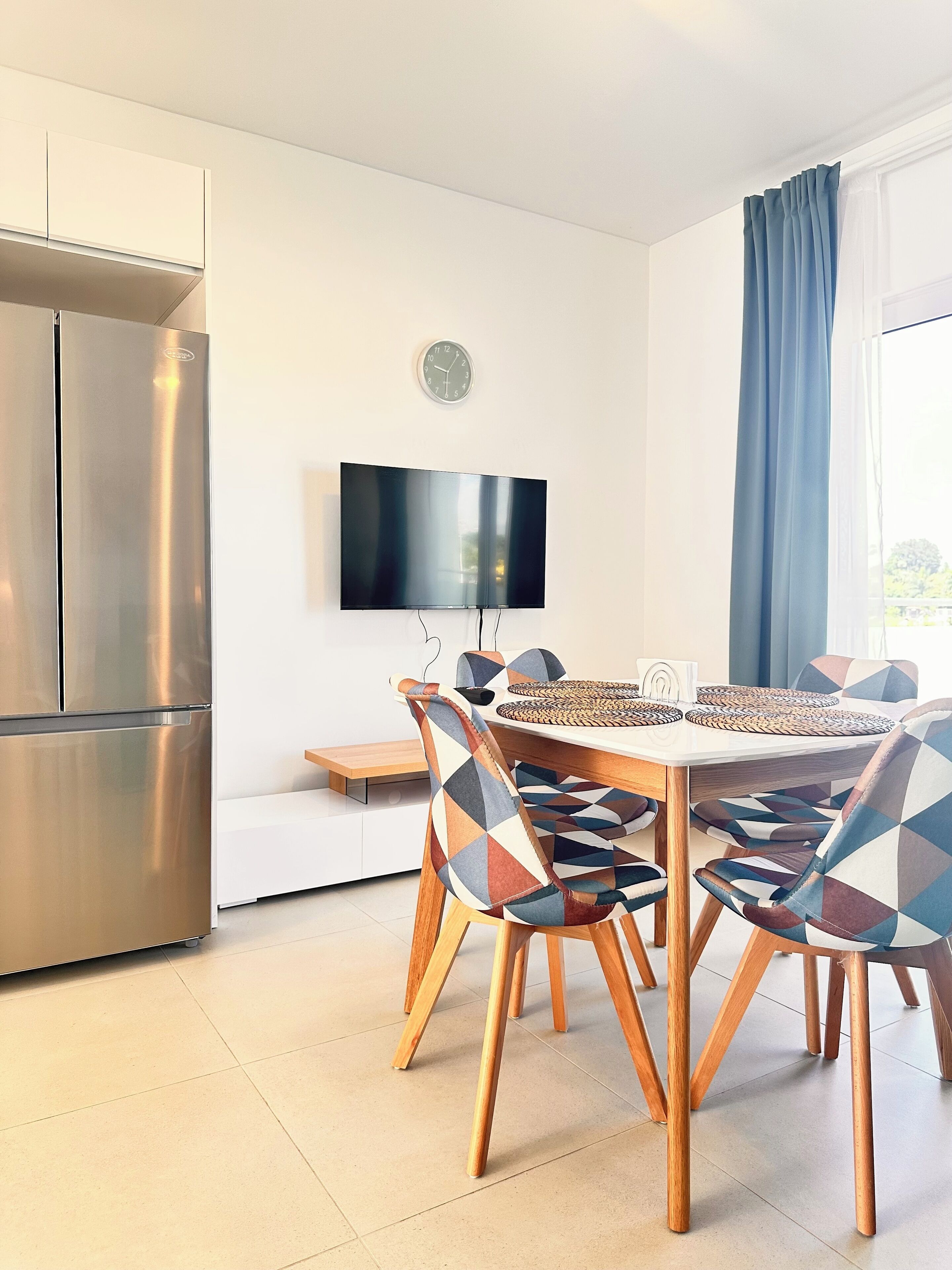 A modern kitchen with a built-in refrigerator, a flat-screen TV mounted on the wall, and a dining area with a wooden table and patterned chairs.