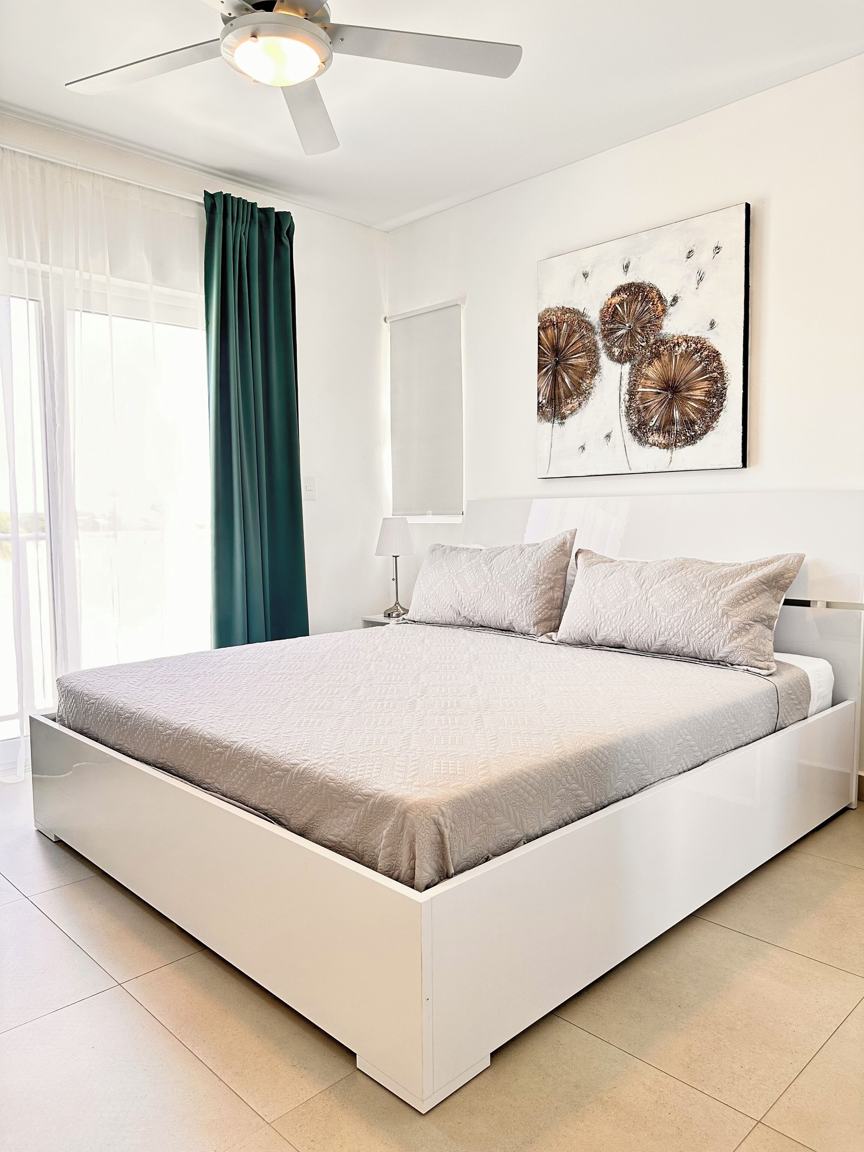 A modern bedroom with a bed, a ceiling fan, a window with curtains, and a wall-mounted artwork.