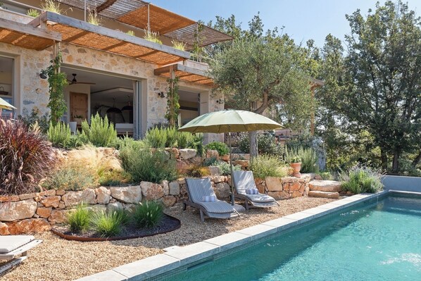 Un espace aménagé au bord de la piscine, avec un mur en pierre, des chaises longues et un parasol.