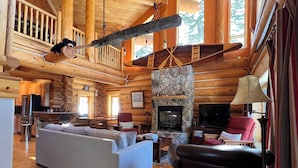 A cozy log cabin interior with a stone fireplace, wooden furniture, and a large window overlooking the outdoors.