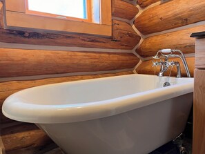 Una bañera blanca en un baño de cabaña de troncos, con paredes de madera y un ventanal.