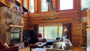 A log cabin interior with a stone fireplace, wooden furniture, and large windows.